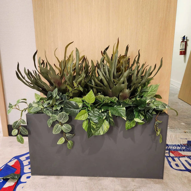 Jardinière artificielle ByNature avec feuillage exotique dense dans un bac rectangulaire graphite devant un mur en bois clair