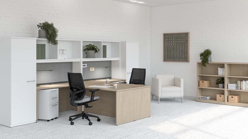 Espace de bureau moderne et lumineux au design épuré. Le mobilier est en bois clair et blanc, avec un grand bureau en L, deux chaises ergonomiques noires, des armoires de rangement et des étagères assorties.