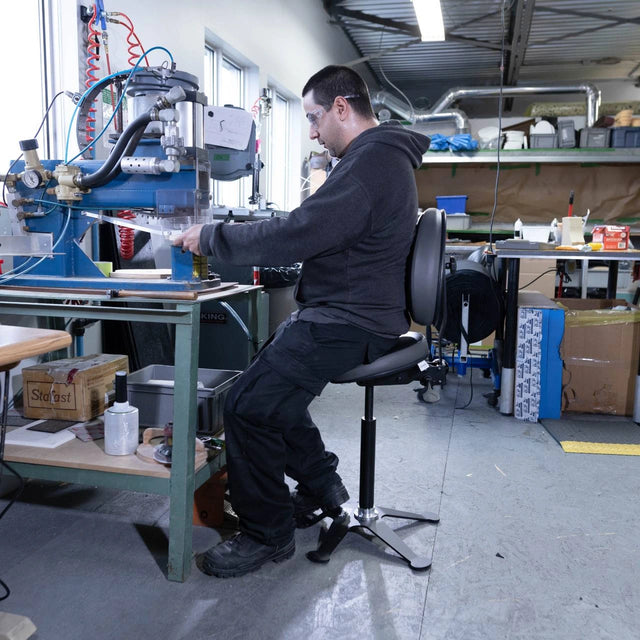 Personne utilisant un siège assis-debout Synetik TA190 en vinyle devant une machine dans un atelier.