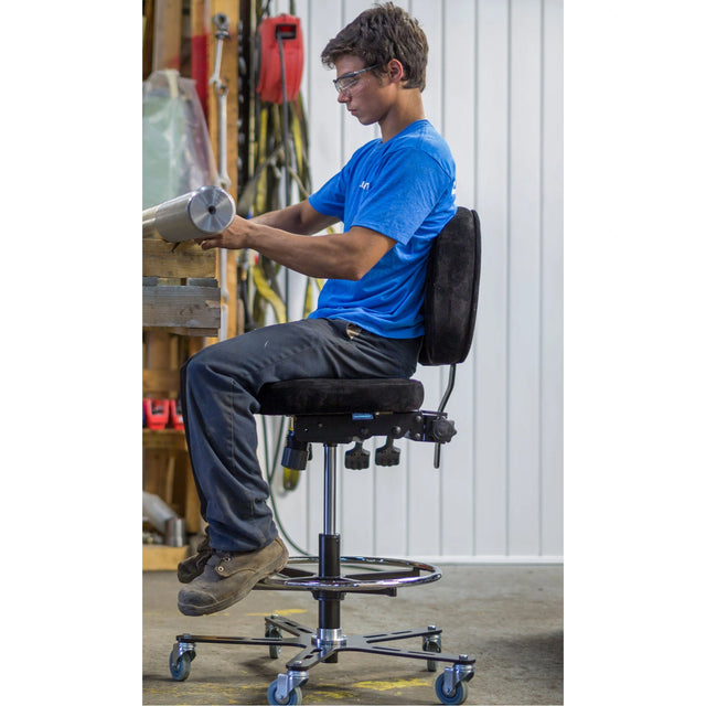 Personne assise sur une chaise haute Synetik TF180 Soudure, les pieds sur l’anneau repose-pieds, devant un établi.